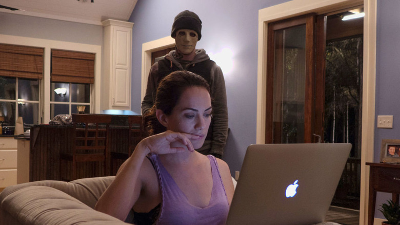 A masked killer stands behind a woman on a laptop