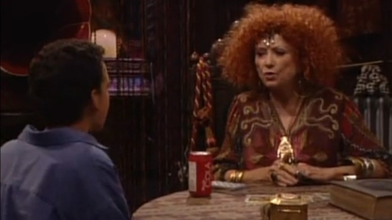 Cory sits opposite Phyllis Diller's fortune teller, dressed in gold with big red hair, at a table in Boy Meets World