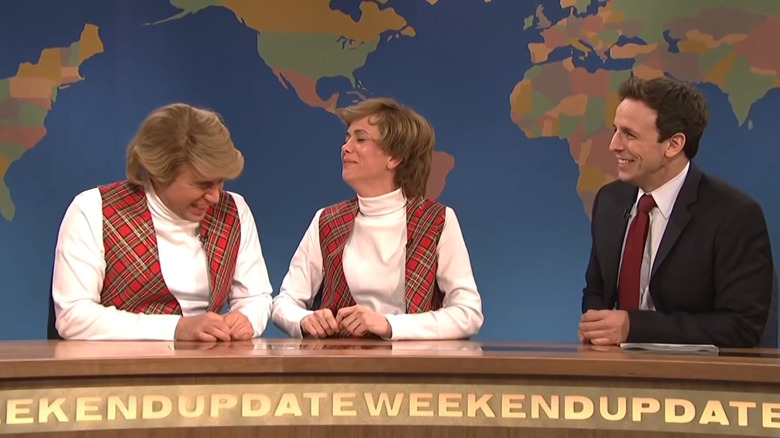 Fred Armisen and Kristen Wiig laughing in white turtlenecks and red vests next to Seth Meyers