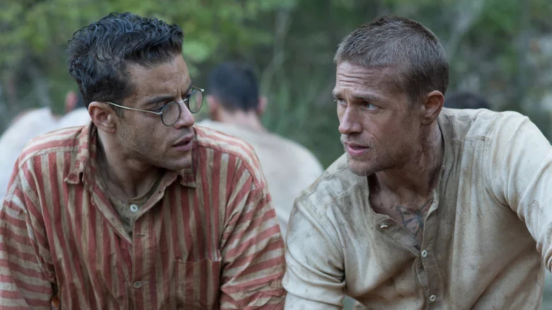 Rami Malek in striped red shirt sitting next to Charlie Hunnanm in Papillon