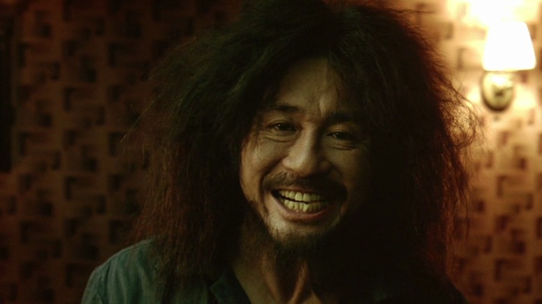 A man with long wild hair smiles creepily at the camera.