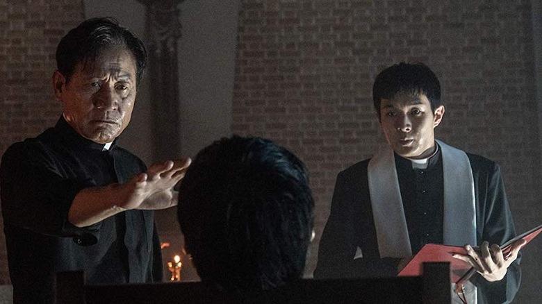 Two priests stand in front of a man sitting in a chair with his back to the camera.