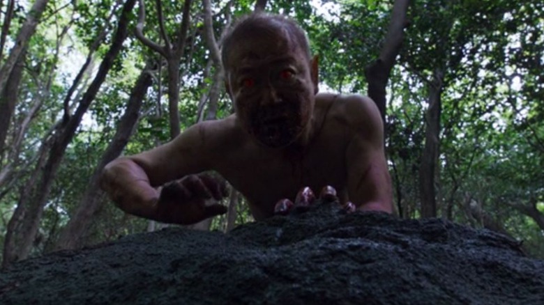 A naked older man with red eyes climbs over a rock.