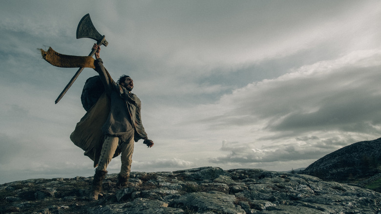 Gawain (Dev Patel) yells while holding axe aloft in The Green Knight