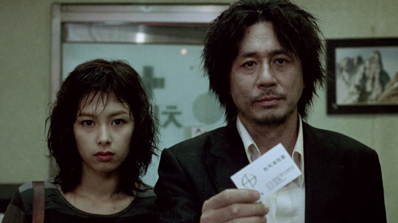 Choi Min-sik as Oh Dae-su standing next to Kang Hye-jung as Mi-do holding business card in Oldboy.