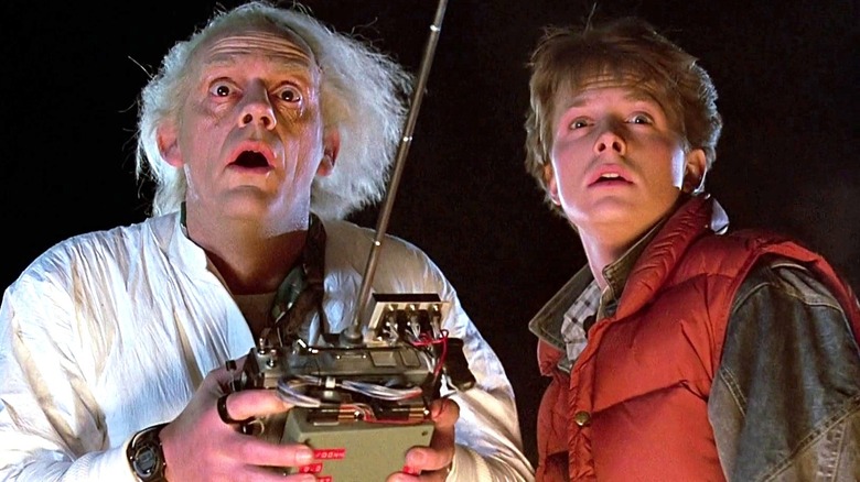 Christopher Lloyd as Dr. Emmett Brown holds a remote control while wearing a white jumpsuit and looks ahead while Michael J. Fox stands next to him as Marty McFly while looking ahead and wearing a red puffer vest and jean jacket in Back to the Future