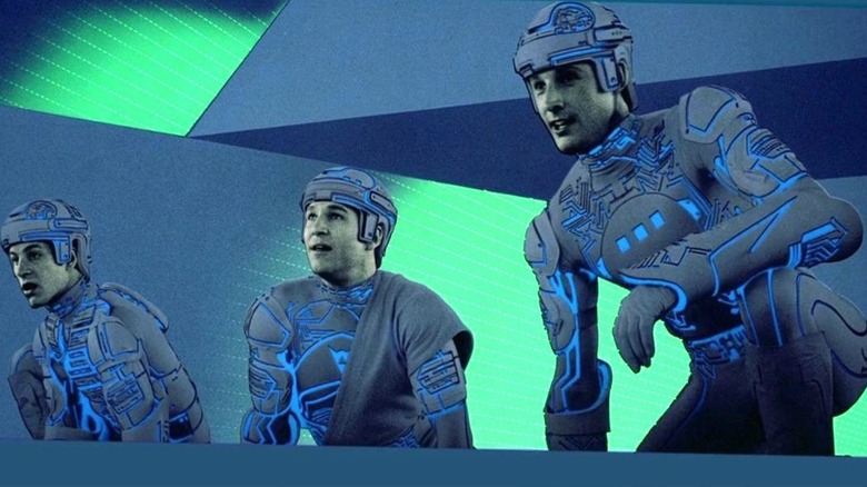 Dan Shor as Ram, Jeff Bridges as Kevin Flynn, and Bruce Boxleitner as Tron all crouch down together in racing suits with a blue and green background behind them in TRON