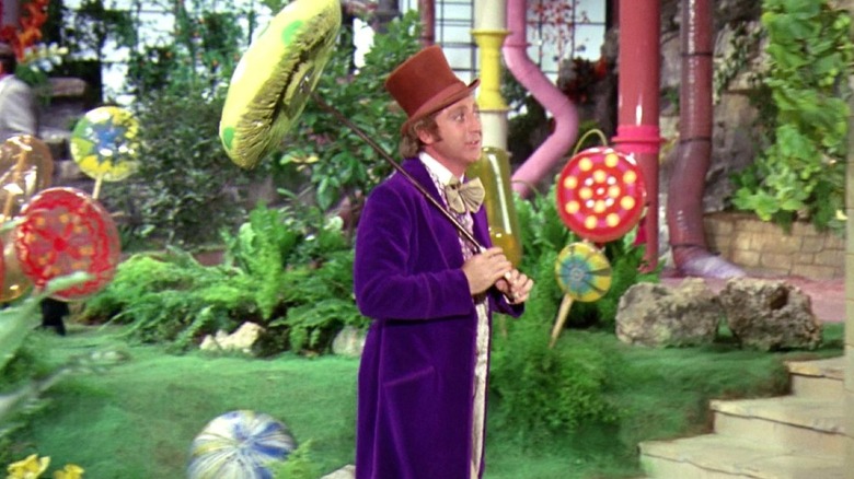 Gene Wilder as Willy Wonka wears a purple suit and brown top hat while holding a mushroom-like parasol while walking through a a garden in his factory in Willy Wonka & the Chocolate Factory