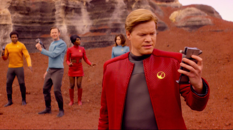 Jesse Plemons as Robert Daly looks down at a scanning device while Osy Ikhile as Nate Packer, Jimmi Simpson as Walton, Michaela Coel as Shania, and Cristin Milioti as Nanette Cole stand behind him surrounded by red rock on Black Mirror