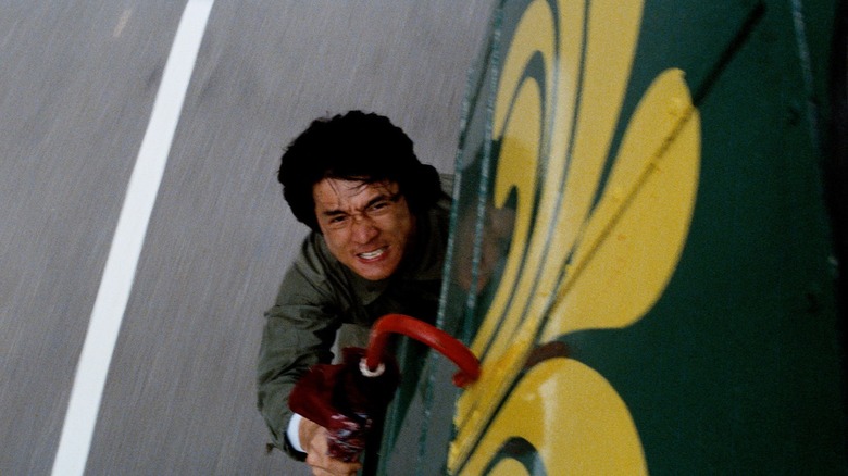 Jackie Chan holding onto the back of a bus with an umbrella