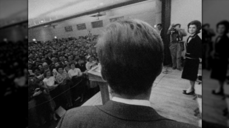 Closeup of JFK on stage, Jackie smiling from right