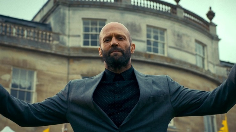 Jason Statham as Adam Clay holding his hands up in front of a mansion in The Beekeeper