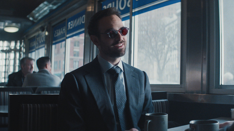 Matt Murdock, wearing a suit and red glasses, sits in a diner booth in Daredevil: Born Again