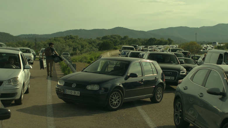 Manel walks down a highway packed with abandoned cars with mountains in the distance