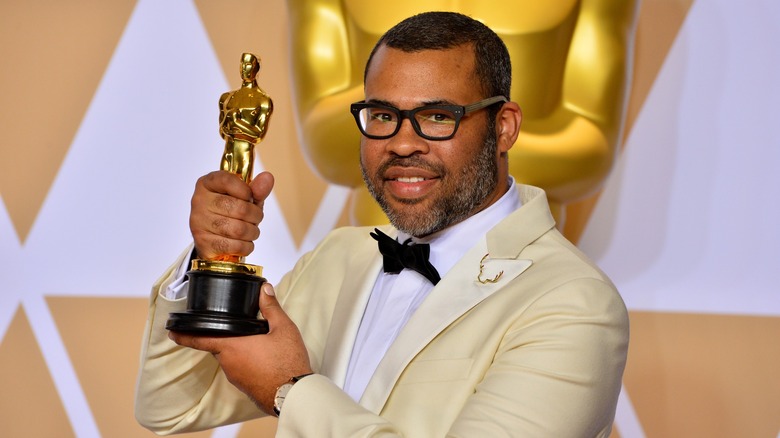 Jordan Peele holding his Oscar up, supporting it by the base