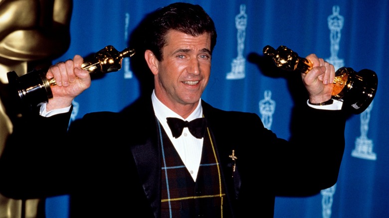 Mel Gibson wearing a tartan vest hoisting two Oscars up above his head