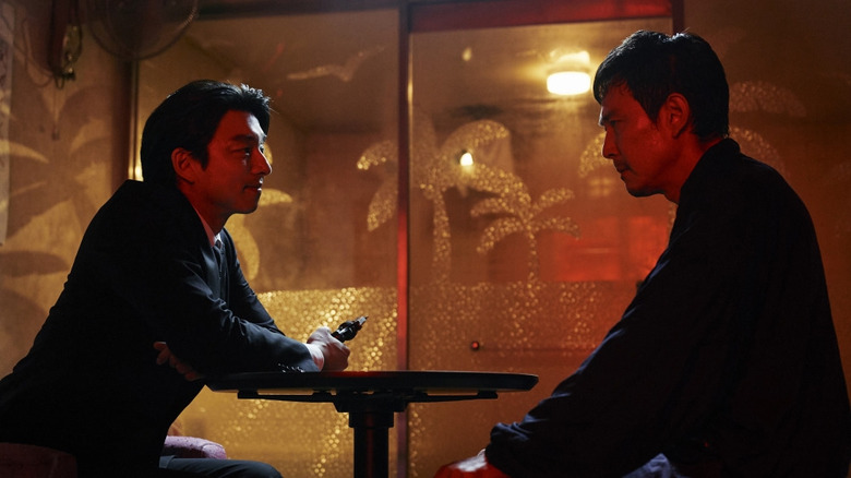 The Recruiter, holding a gun, sits at a table with Seong Gi-hun