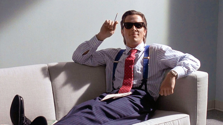 Patrick Bateman wears sunglasses and sits on a couch