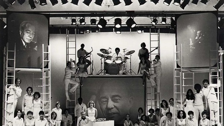 A group of young people on a stage, including a handful playing musical instruments, with screens showing an old man in three separate poses