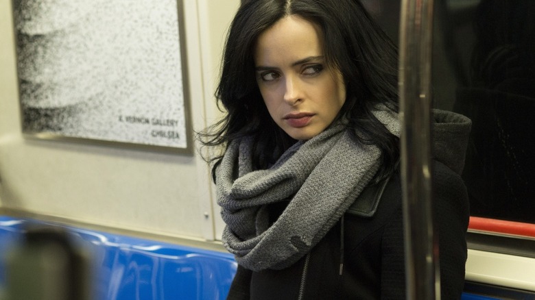 a brunette woman stares to the side while sitting on the subway in a black jacket and grey scarf