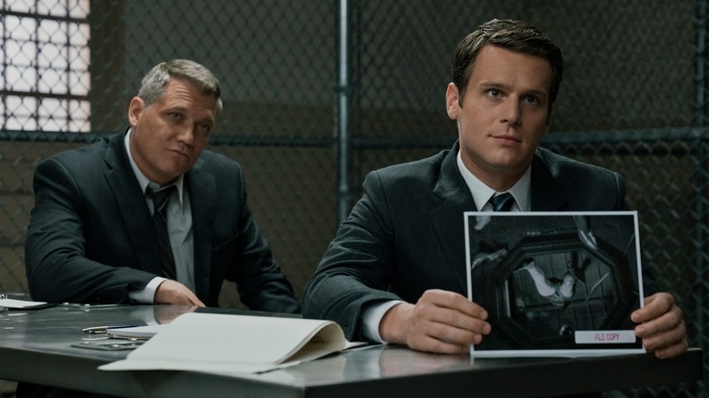 Bill Tench (Holt McCallany) and Holden Ford (Jonathan Groff) in an interrogation room, holding up a crime scene photo on Mindhunter