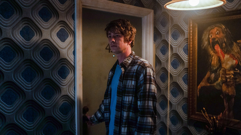 a man in a plaid shirt stands in a hallway with patterned wallpaper and the goya's painting 'saturn devouring his son' on the wall