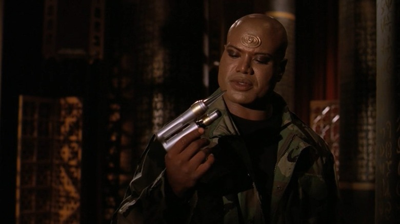 Christopher Juiz Teal'c olhando para uma arma que ele está segurando em Stargate: SG-1