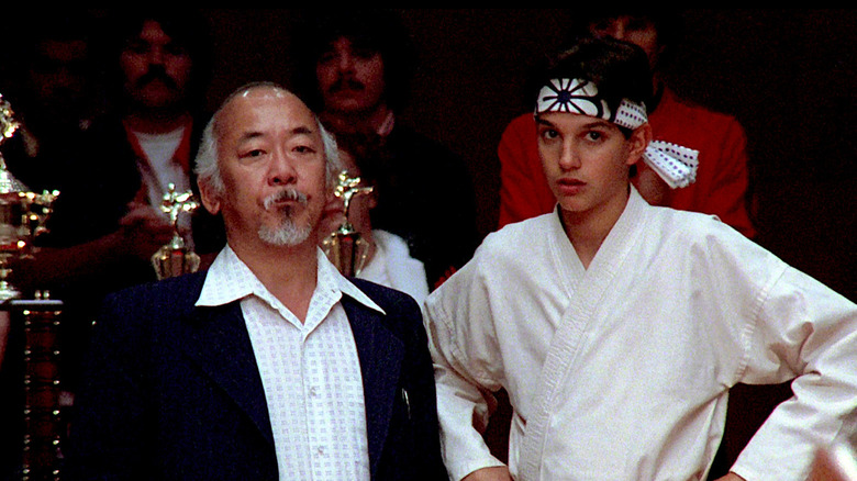 Mr. Miyagi and Daniel standing next to trophies at a karate tournament in The Karate Kid