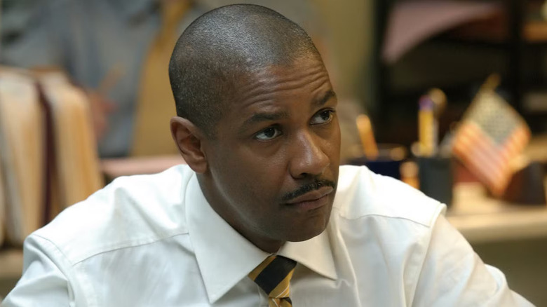 Denzel Washington as Detective Keith Frazier, wearing a white-collared shirt and tie, with a focused expression, stands in front of blurry desks in the background from Inside Man (2006).