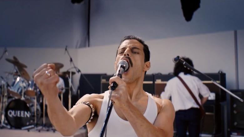 Rami Malek as Freddie Mercury singing on stage with his arm extended in front of him in Bohemian Rhapsody