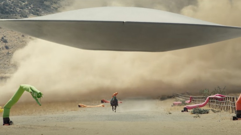 a flying saucer low to the ground surrounded by blow up zippy men and a horse riding below it in the movie nope