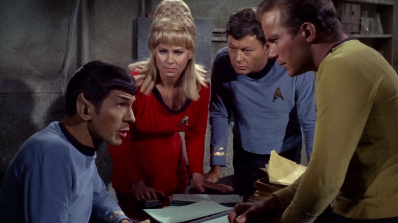 Spock seated at an old, 20th century table with Yeoman Rand, McCoy, and Kirk standing over him in Star Trek