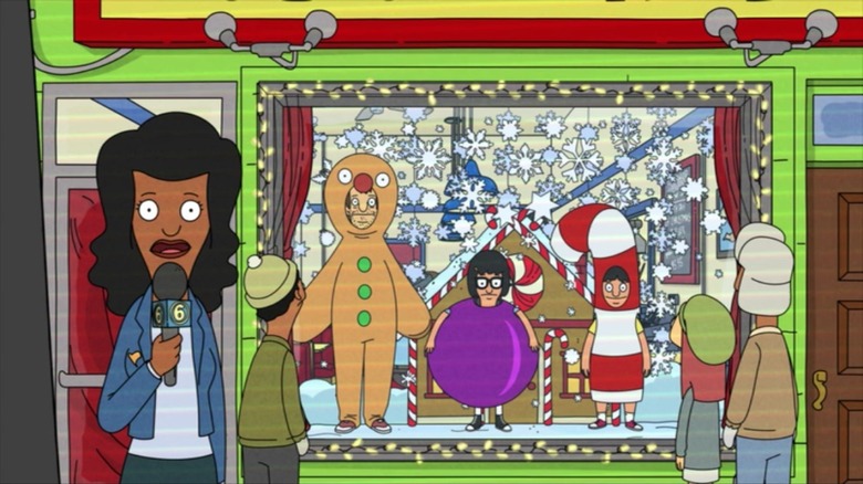 A news reporter stands outside Bob's Burgers, where there is a festive window display