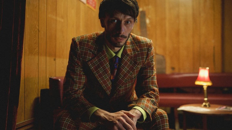 Richard Gadd's Donny sits in an empty room, looking haunted, in Baby Reindeer. He's also wearing a clashing plaid suit.