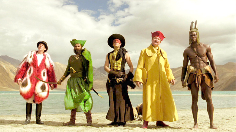 Five men dressed in colorful outfits stand in a desert oasis