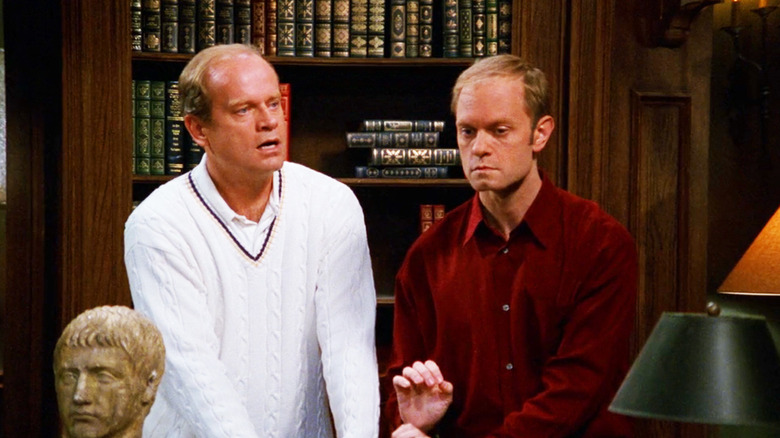 Frasier e Niles estão em frente a uma estante de livros em uma biblioteca