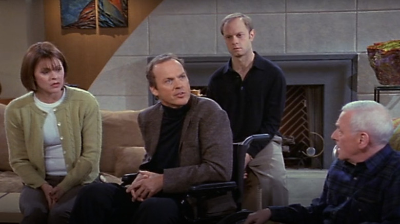 Michael Keaton as Blaine Sternin seated in a wheelchair gesturing aggressively with Jane Leeves as Daphne Moon, David Hyde Pierce as Niles Crane, and John Mahoney as Martin Crane sitting around him staring in Frasier.