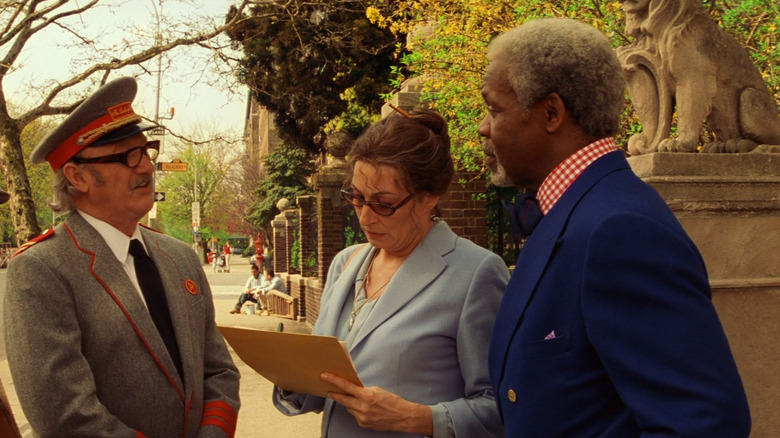 Royal Tenenbaum (Gene Hackman), d﻿ressed as a hotel elevato﻿r ope﻿rato﻿r, talks to Etheline Tenenbaum (Anjelica Huston) and Hen﻿ry She﻿rman (Danny Glove﻿r) on a New Yo﻿rk sidewalk