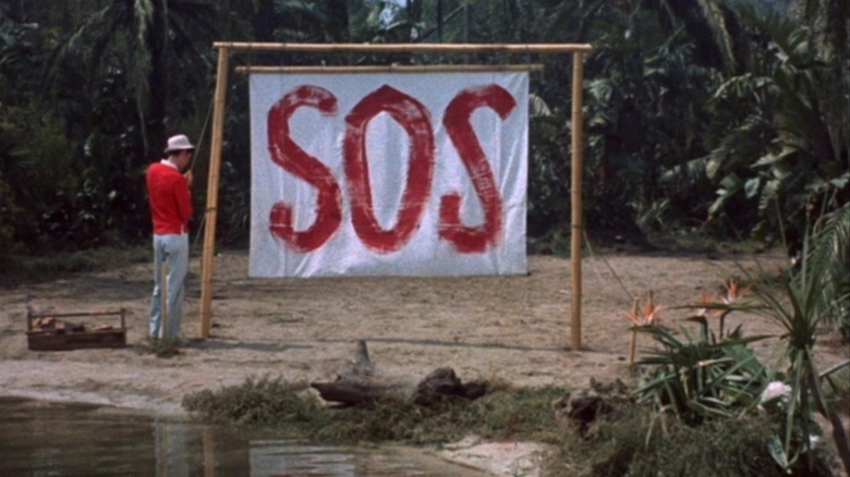 Gilligan standing next to a big S.O.S. banner on the lagoon beach in Gilligan's Island