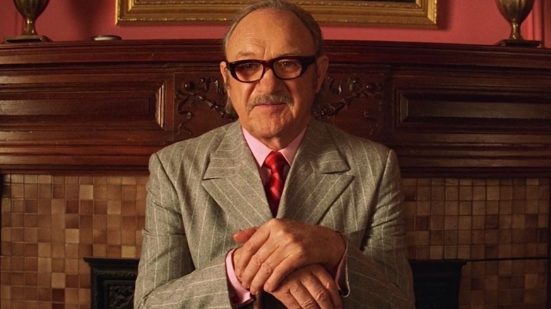 Royal Tenenbaum, wearing a dapper suit and tie, smiles enigmatically and sits in front of a credenza