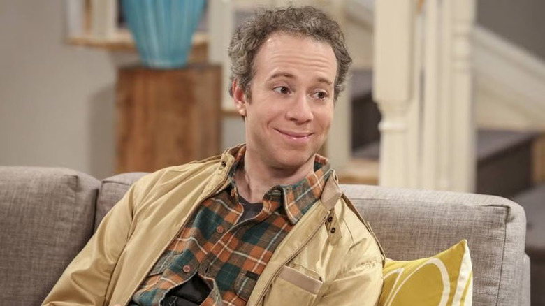 Stuart with gray hair and a yellow shirt sitting on a couch in The Big Bang Theory