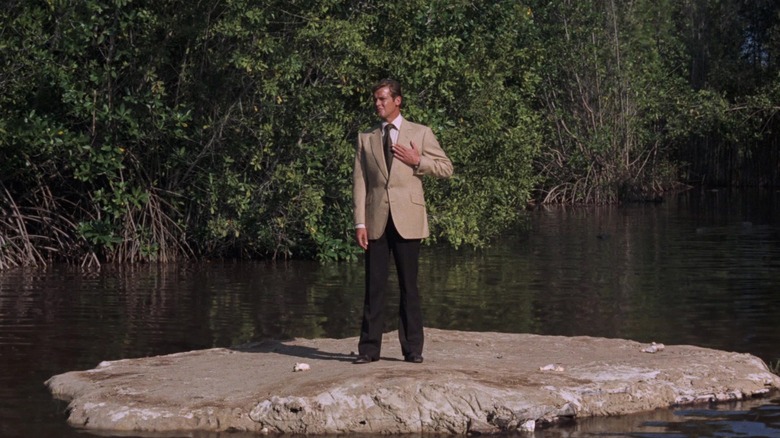 Roger Moore's James Bond stands in a tan suit on a small piece on land in the middle of a lake in Live and Let Die