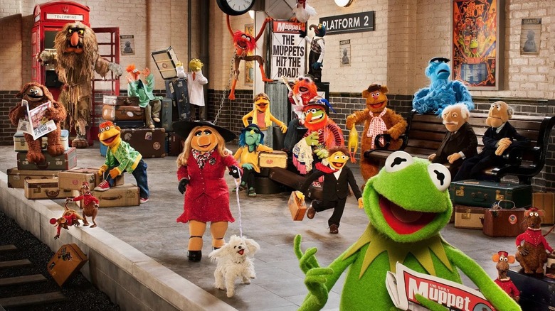 On a train platform, a bevy of Muppets (including Scooter, Sweetums, Miss Piggy, Kermit the Frog, and Fozzie Bear) look at the camera with big smiles on their faces