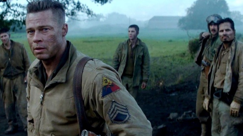 Norman stands in a field wearing a military jacket, looking serious and worn-out, with the rest of the ensemble in the background in Fury