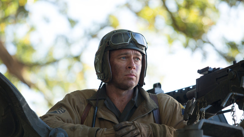 Don has his gloved hands clasped, a pilot's helmet with goggles resting on his head, looking into the distance pensively next to a machine gun in Fury