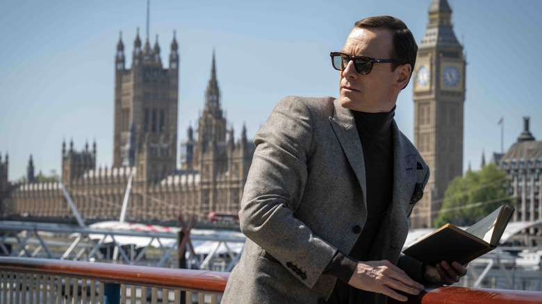 Michael Fassbender as George Woodhouse standing on a bridge with a book in Black Bag