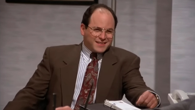 George Costanza in a brown suit, sitting opposite his boss' desk.