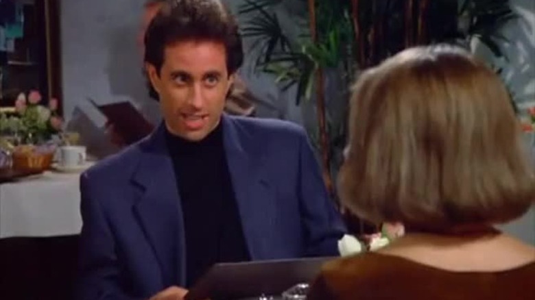 Seinfeld at the dining table, opening the menu, looks suspiciously at his date. Scene from Seinfeld.