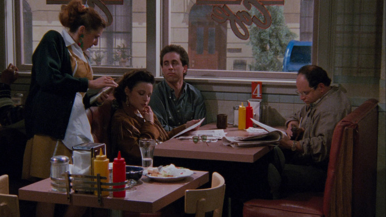 Julia Louis-Dreyfus as Elaine, Jerry Seinfeld as Jerry, and Jason Alexander as George sitting at a restaurant booth on Seinfeld