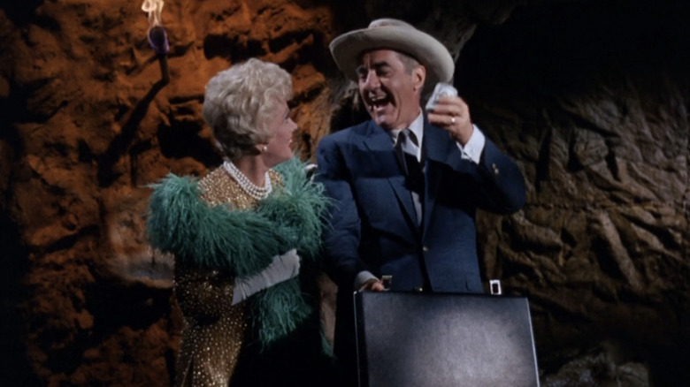 Natalie Schafer's Mrs. Howell berdiri bersama Jim Backus 'Thurston Howell III saat ia mengangkat uang tunai dengan penuh kemenangan di Pulau Gilligan